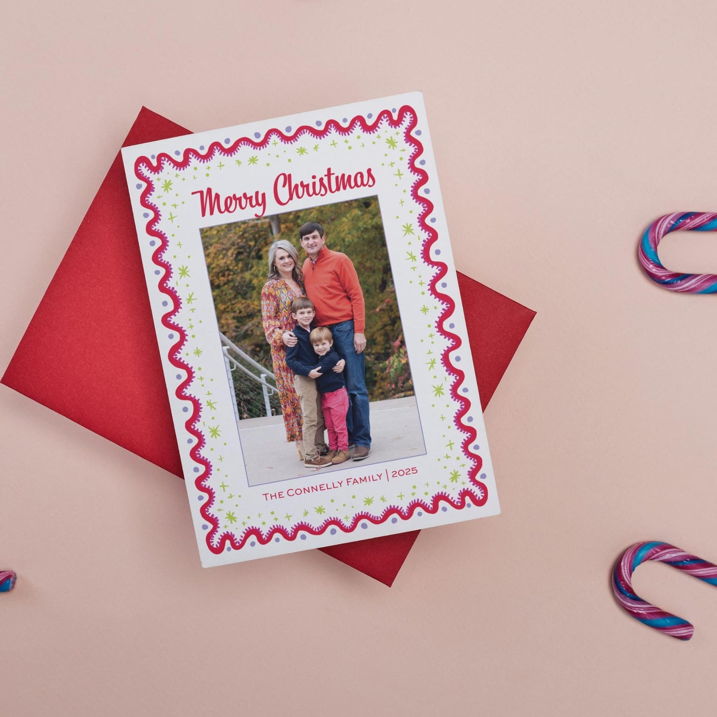 A family portrait of a man, woman, and child is displayed on a red card with a festive border, placed on a pink background with candy canes.