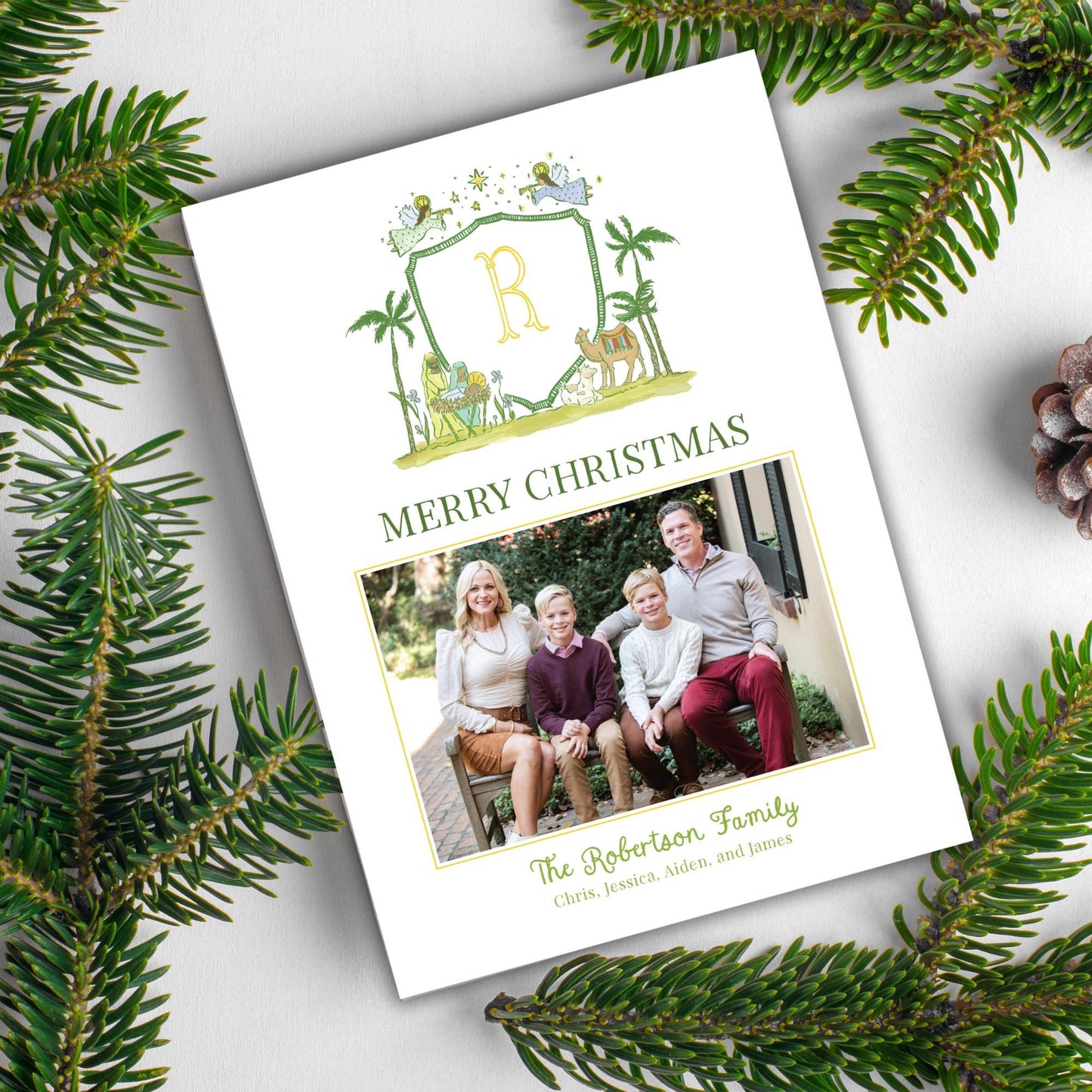 A white card with a family photo and a crest design, surrounded by pine branches and pine cones, indicating a holiday or seasonal setting.