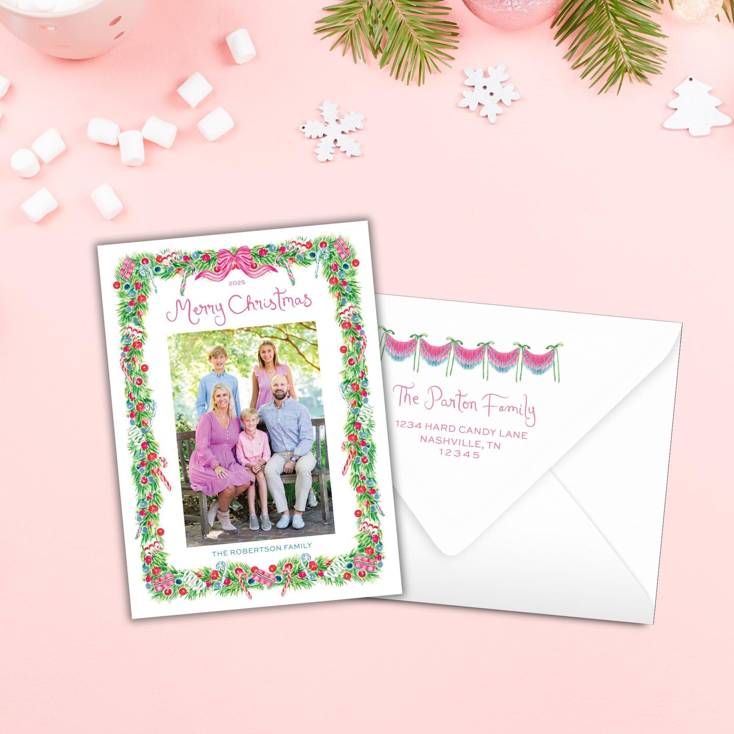 A Christmas card with a family photo and a white envelope with a personalized address, both placed on a pink background with snowflakes and other holiday decorations.