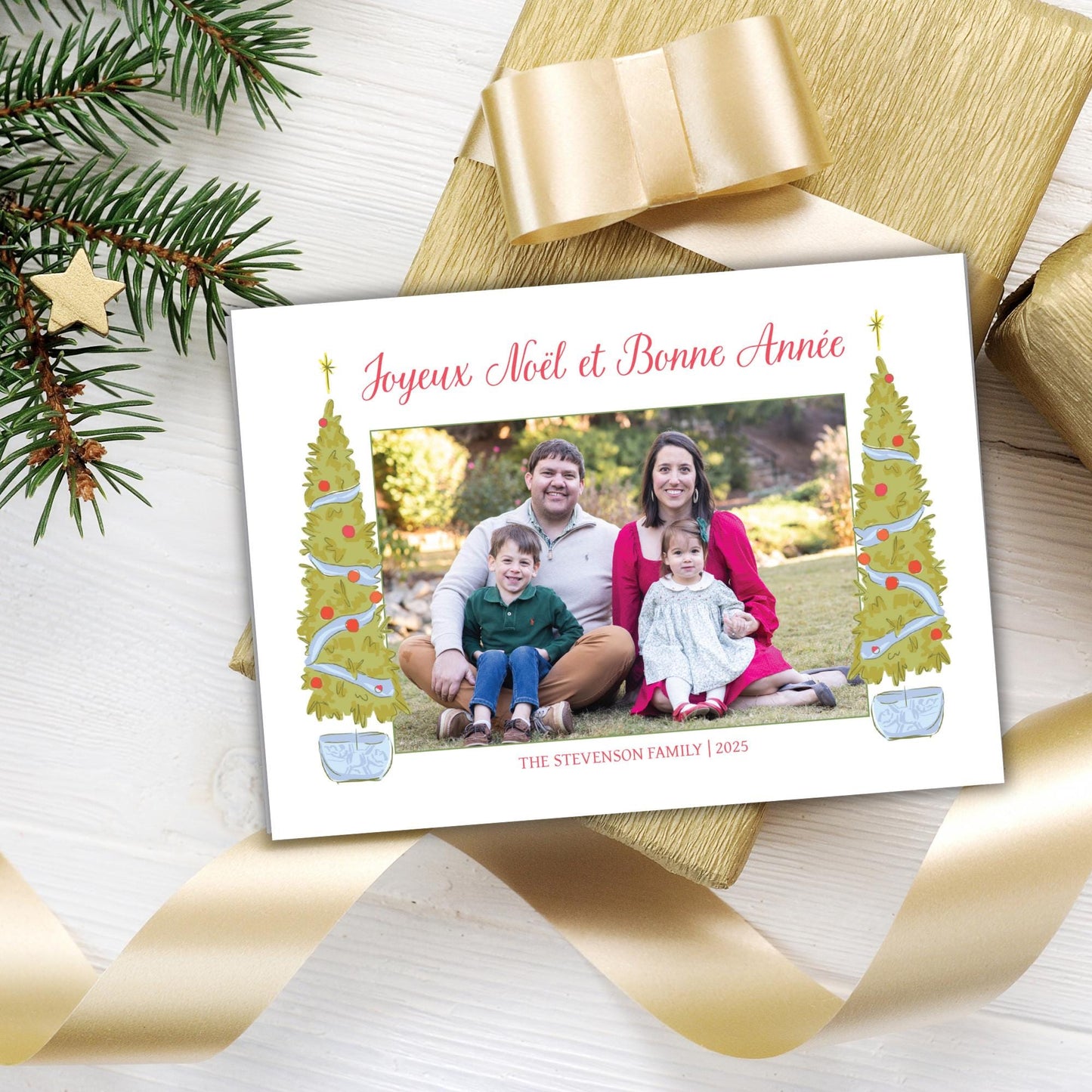 A family portrait of a man, woman, and two children sitting together on a grassy area, surrounded by Christmas trees and a ribbon.