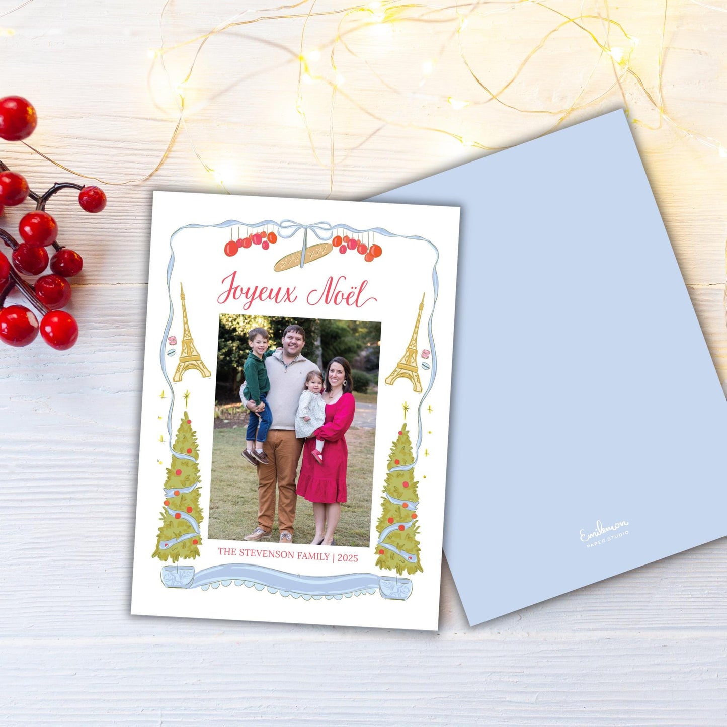 A family photo is displayed on a card with the text "Joyeux Noel" and "The Steveston Family 2015" printed on it. The card is placed on a wooden surface, with a string of lights and red berries in the background.