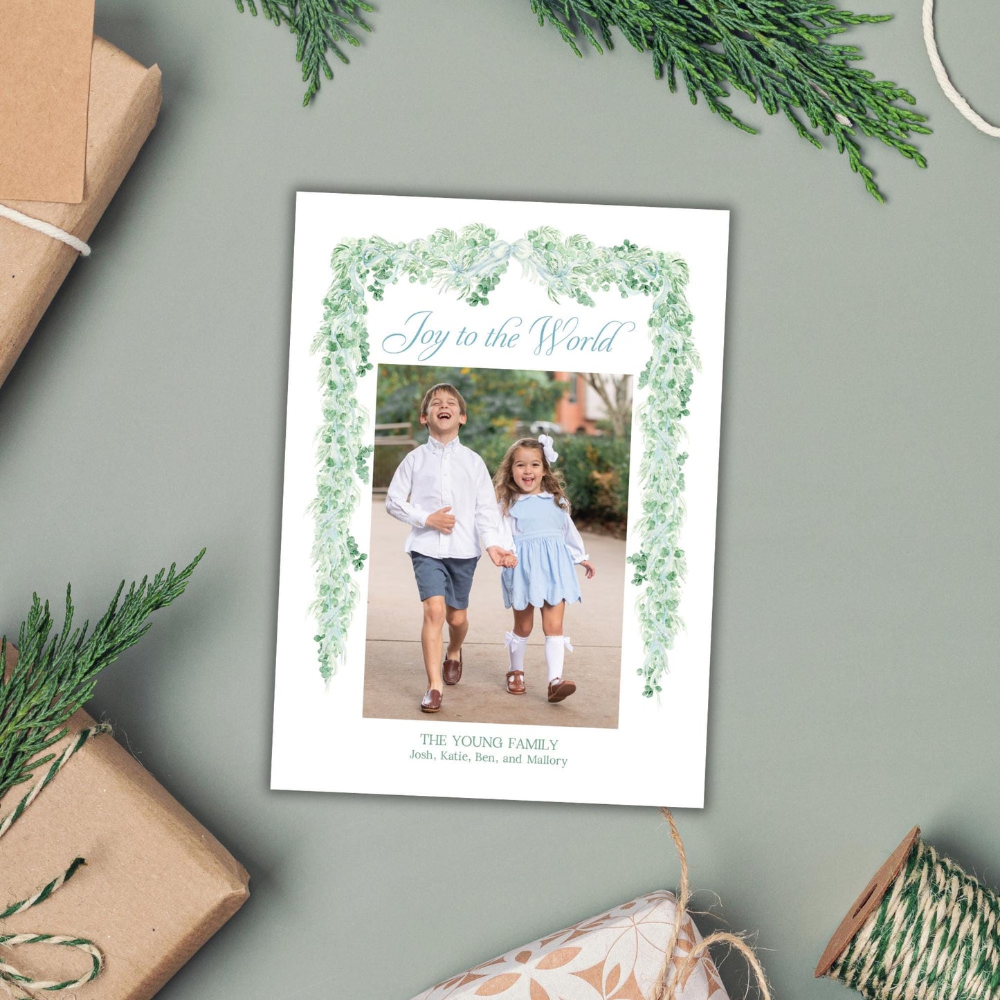 A white card with a photo of two children, surrounded by greenery and holiday decorations, including wrapped gifts and sprigs of pine.
