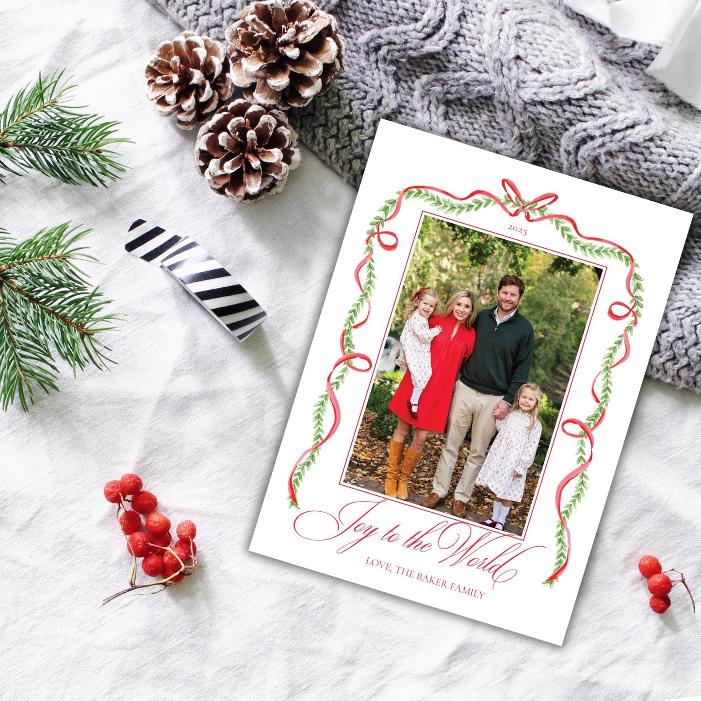A Christmas card with a family photo is placed on a white cloth, surrounded by pine branches, pinecones, and red berries.