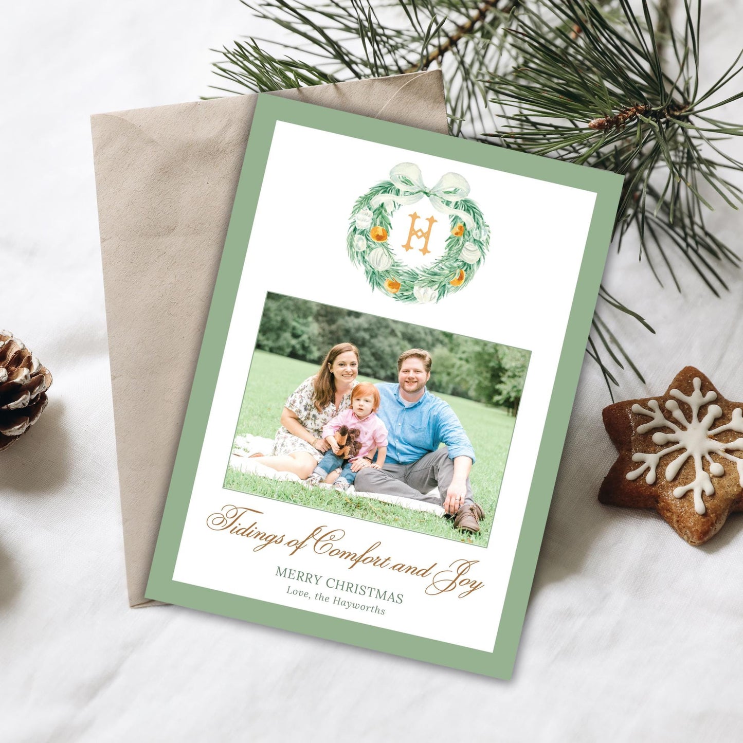 A Christmas card with a family photo and a wreath, along with some pine cones and a star-shaped cookie, all placed on a white surface.