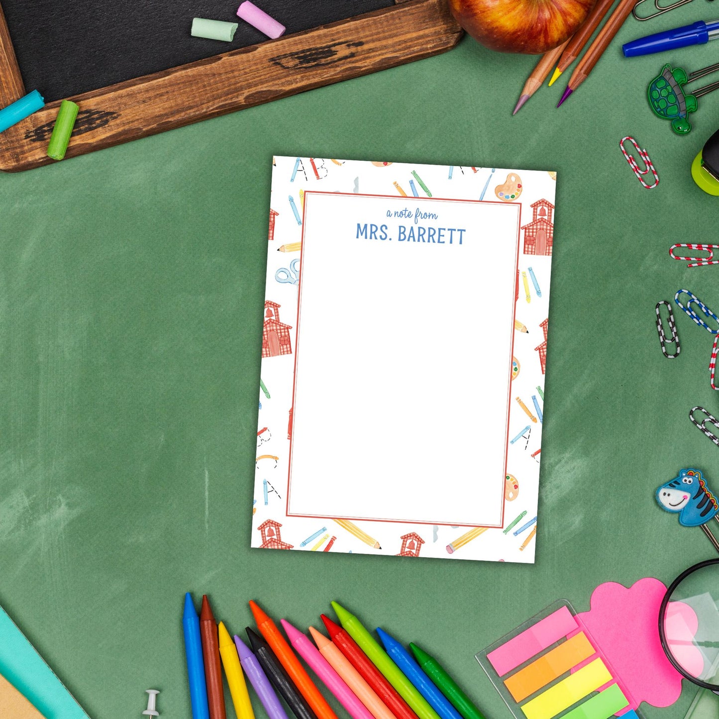 A blank personalized note card with a name and a school-themed design, surrounded by various school supplies such as pencils, markers, and a magnifying glass, all arranged on a green surface.