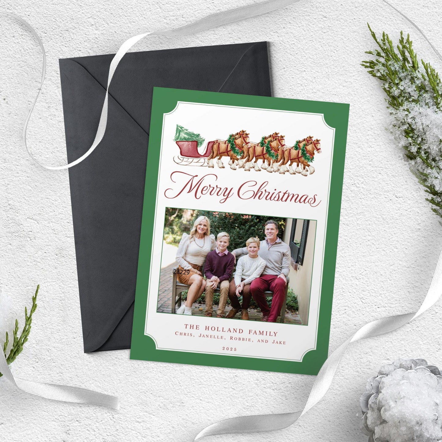 A Christmas card with a family photo and a sleigh with reindeer on it, placed on a white surface with a black envelope and some greenery.