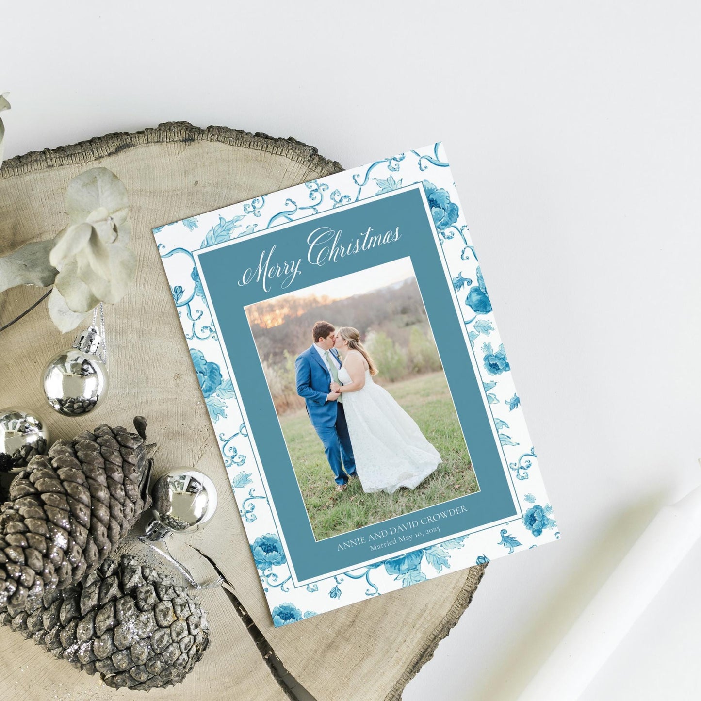 A wedding invitation card with a photo of a couple standing together, surrounded by holiday decorations such as pine cones and ornaments.