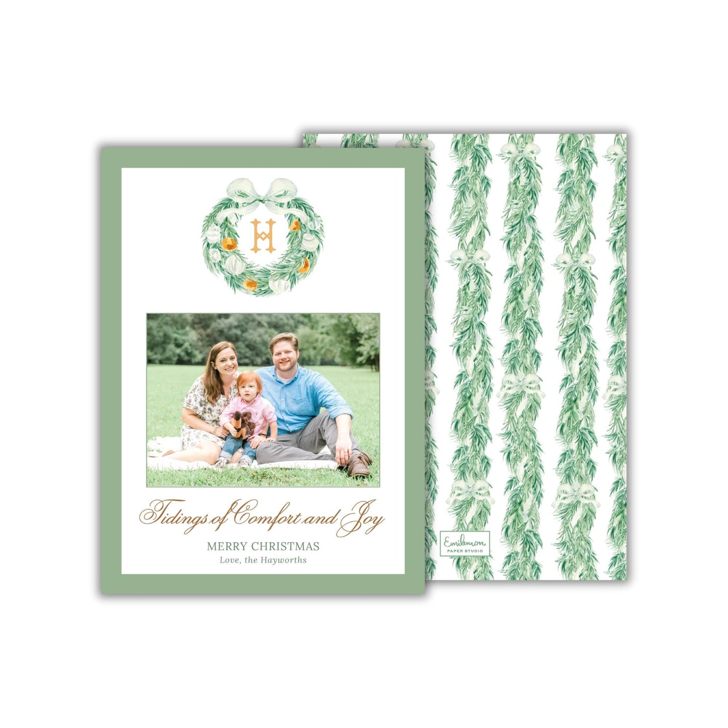 A family portrait of a man, woman, and child sitting together on a blanket in a grassy field, with a wreath above them.