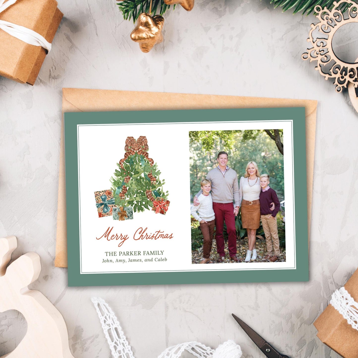 A Christmas card featuring a family photo and a Christmas tree illustration, placed on a white surface with other holiday decorations and gifts.