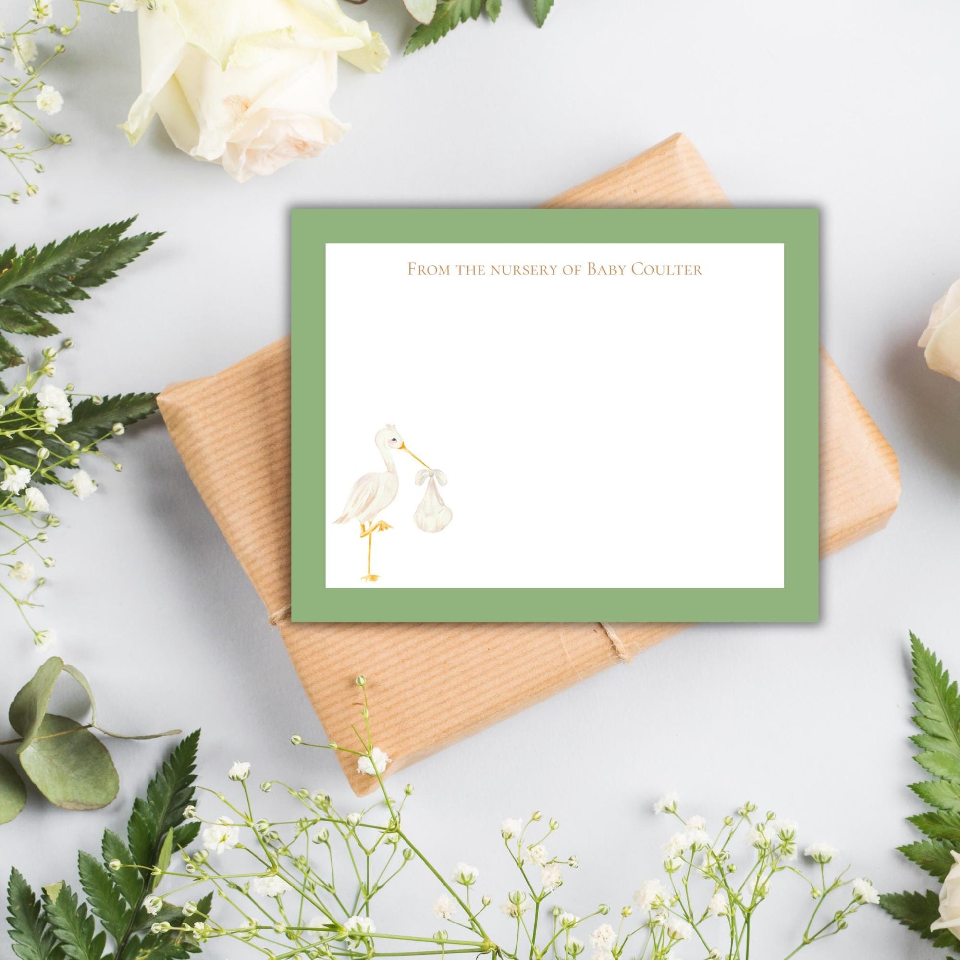 a green-framed note with a baby stork illustration, placed on a white background with some flowers and leaves.