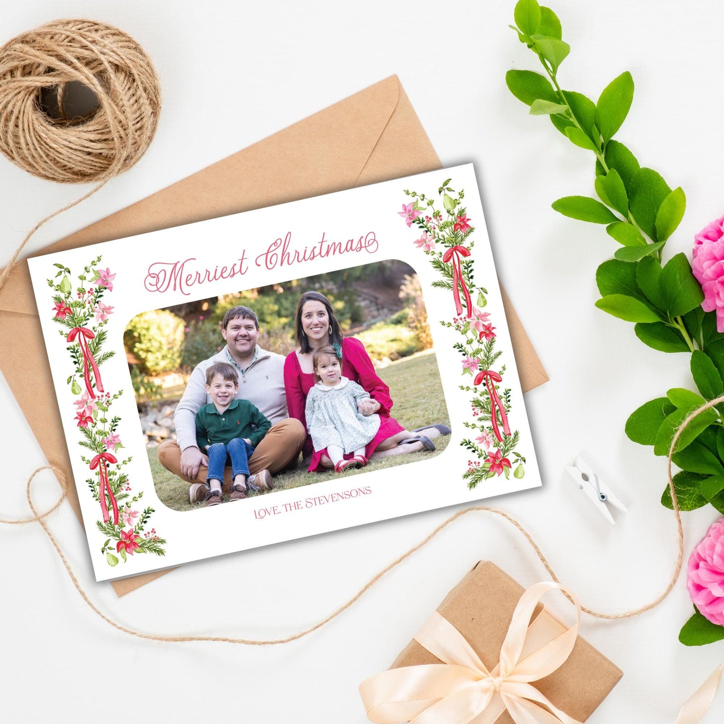 A family photo is displayed in a frame on a white background, surrounded by holiday decorations such as a spool of twine, a gift box, and a bouquet of pink flowers.