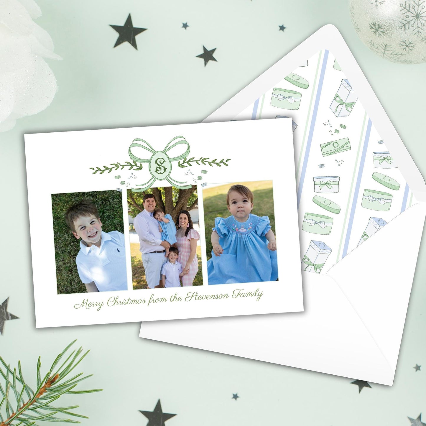 A family photo is displayed on a white card with a green ribbon, accompanied by a matching envelope with a pattern of Christmas-themed illustrations.