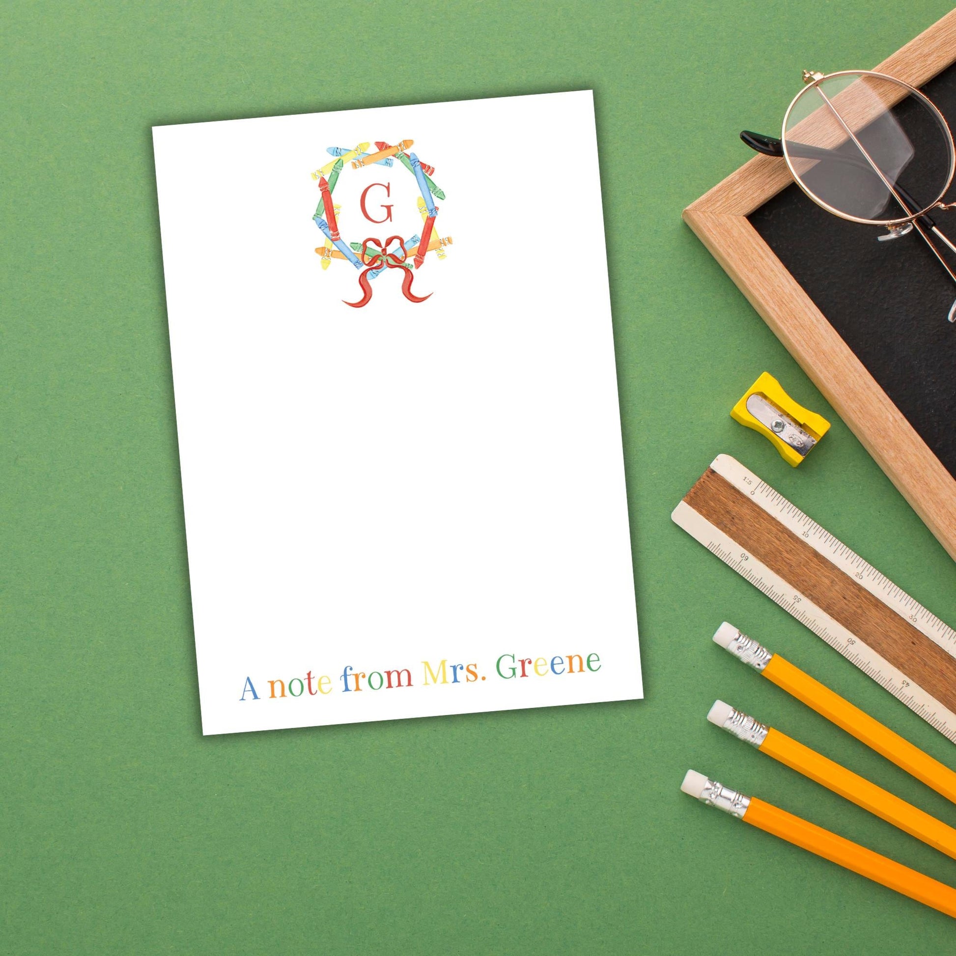 a blank personalized note card with a colorful wreath design and the letter "G" on it, placed on a green surface alongside a pair of glasses, a ruler, and a pencil sharpener.