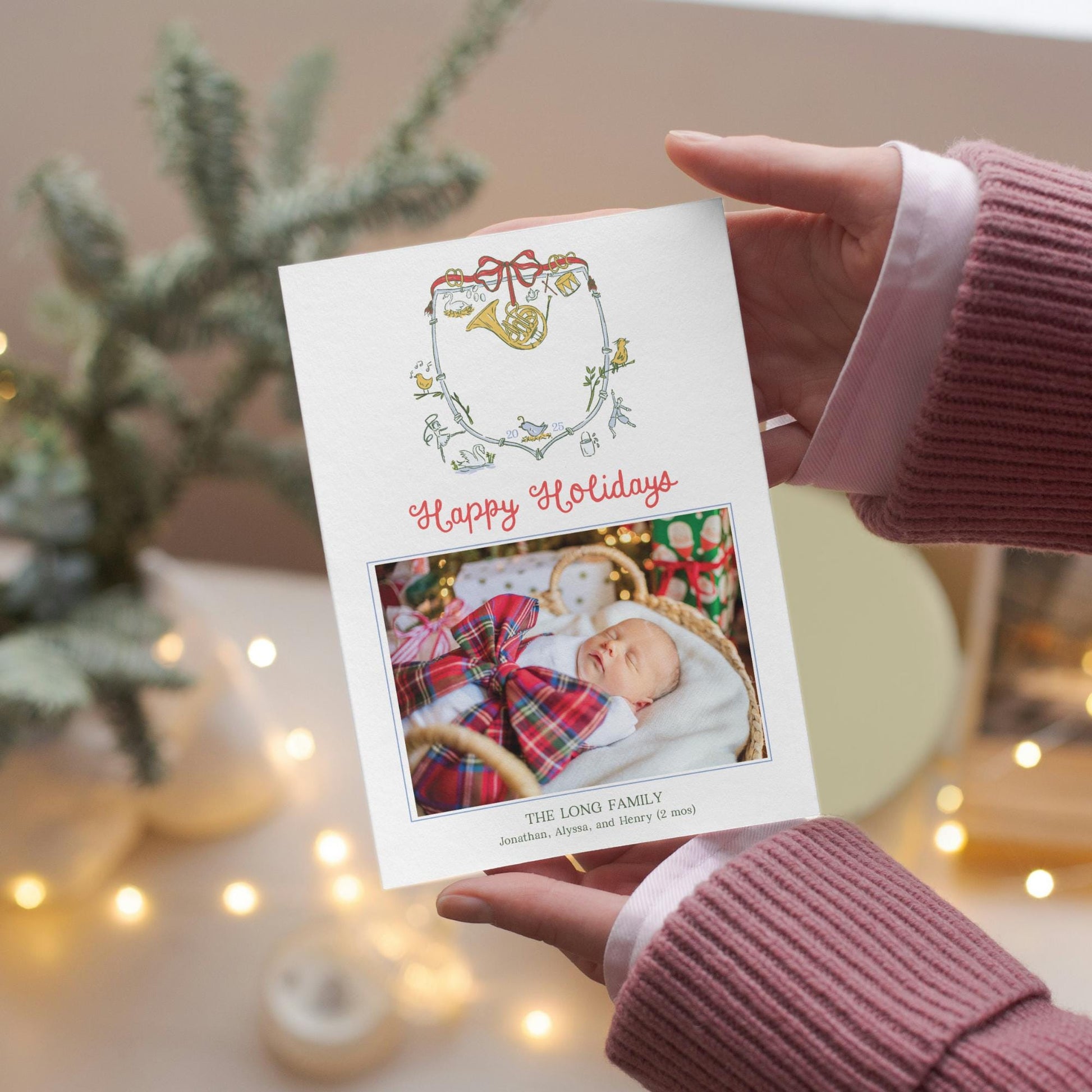 A person&#39;s hands holding a white holiday greeting card with a photo of a baby and the text &quot;Happy Holidays&quot; and &quot;The Long Family&quot; printed on it.
