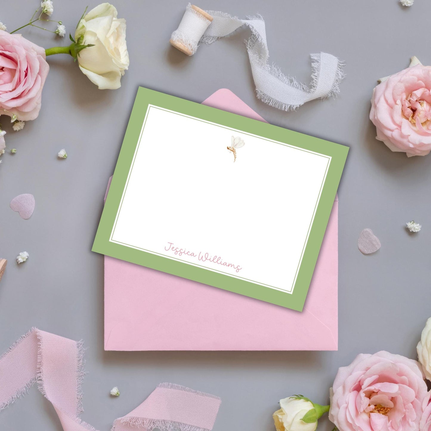 a pink envelope with a green frame, a blank white card with a floral design, and various pink and white flowers scattered around the envelope and card.