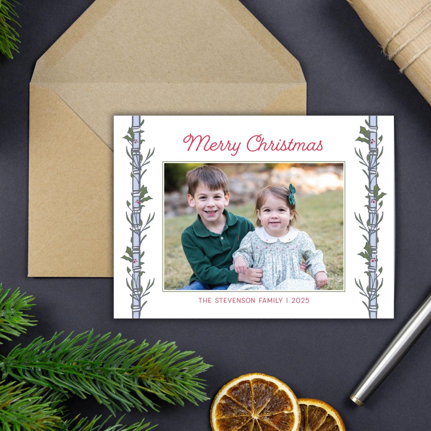 A Christmas card with a photo of two children, a boy and a girl, sitting together in a field. The card has a festive design with a tree branch and greenery on the edges. The card is placed on a dark surface, with a pen and some dried orange slices nearby.