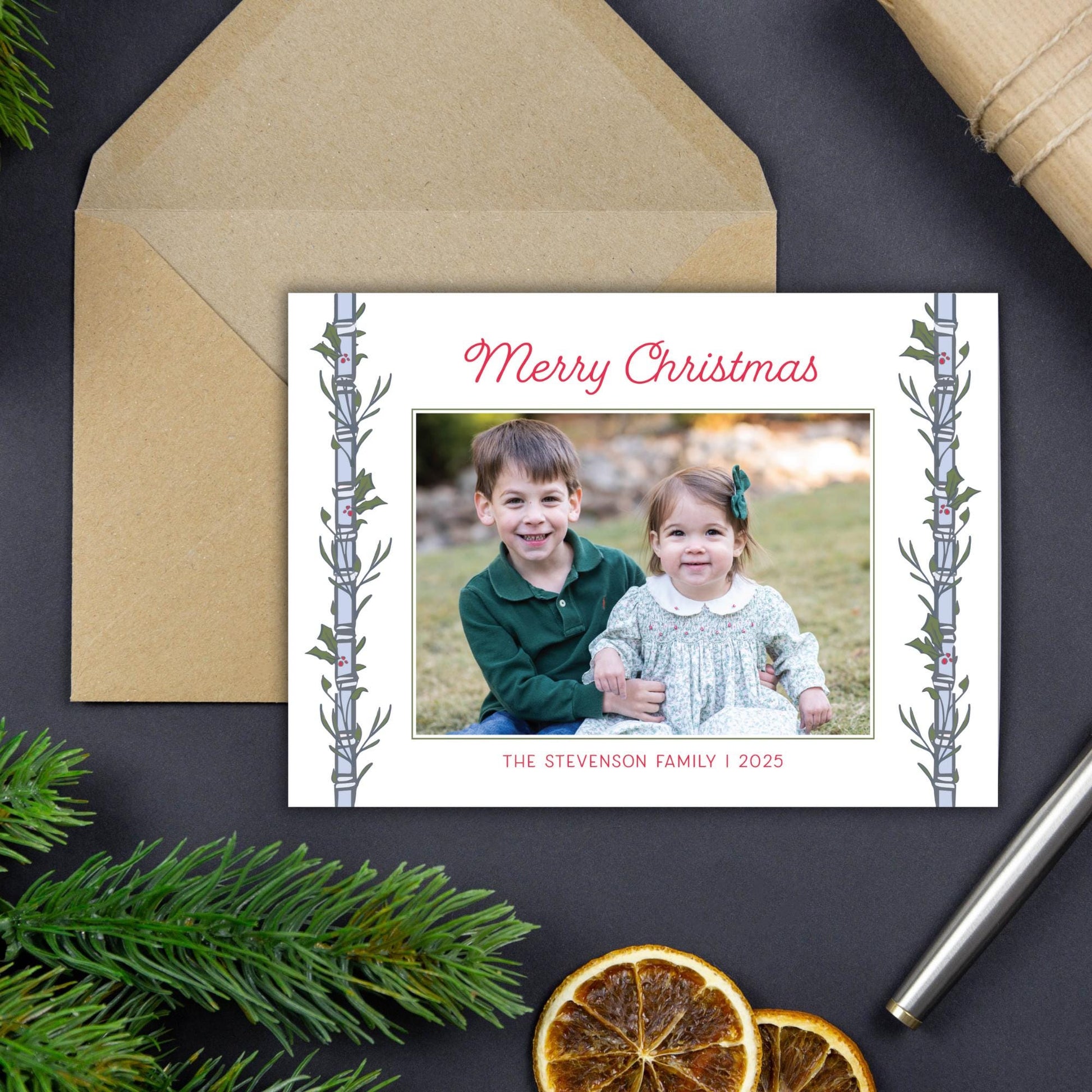 A Christmas card with a photo of two children, a boy and a girl, sitting together in a field. The card has a festive design with a tree branch and greenery on the edges. The card is placed on a dark surface, with a pen and some dried orange slices nearby.