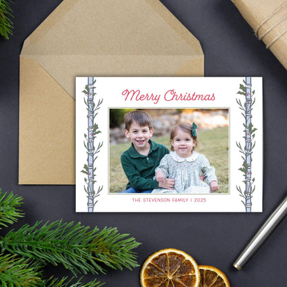 A Christmas card with a photo of two children, a boy and a girl, sitting together in a field. The card has a festive design with a tree branch and greenery on the edges. The card is placed on a dark surface, with a pen and some dried orange slices nearby.