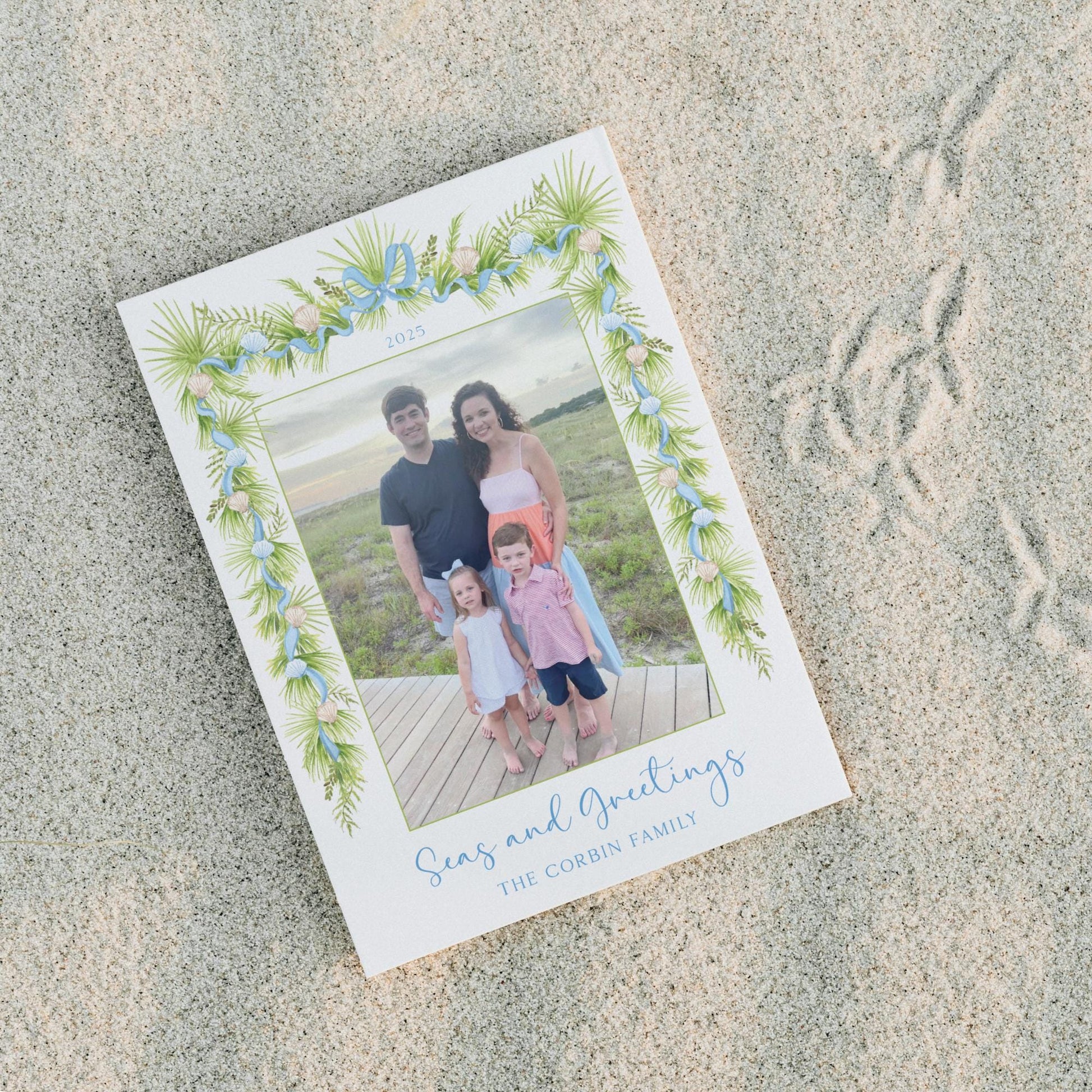 A family photo of a man, woman, and two children, with a decorative border of green leaves and flowers, placed on a sandy beach.