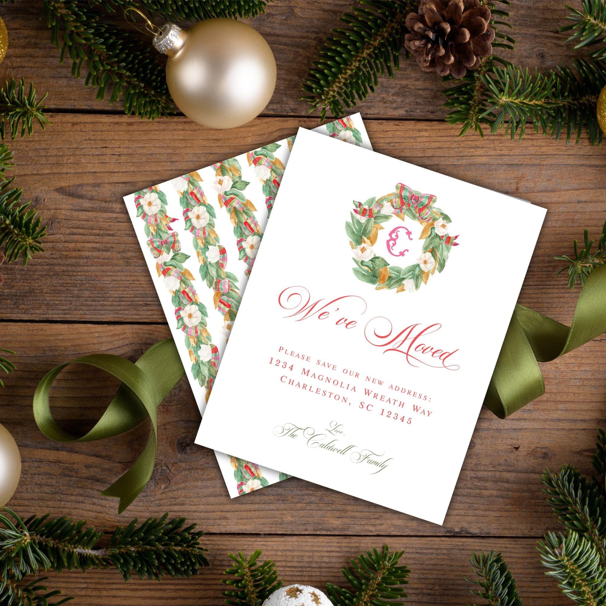 A white card with a wreath design and text is placed on a wooden surface, surrounded by Christmas decorations such as pine branches, ornaments, and a ribbon.
