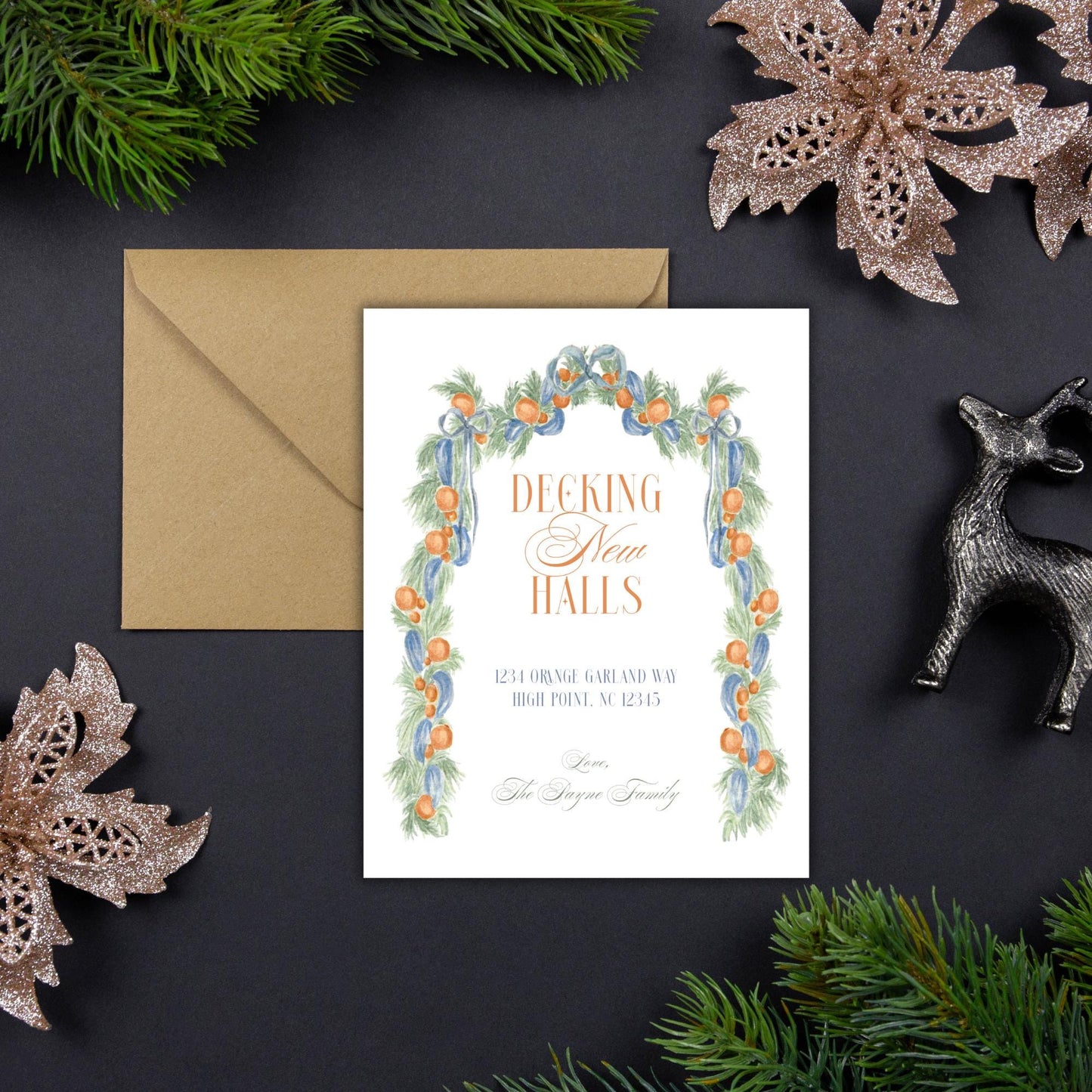 A white card with a decorative border featuring orange and green elements, surrounded by holiday decorations such as pine branches, a silver deer figurine, and a gold star-shaped ornament.