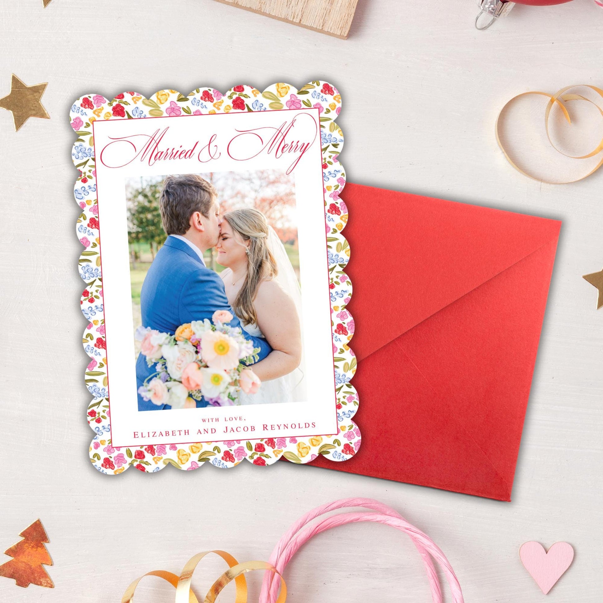 A wedding card with a photo of a couple embracing is placed next to an envelope on a white surface, surrounded by Christmas ornaments and a pink ribbon.