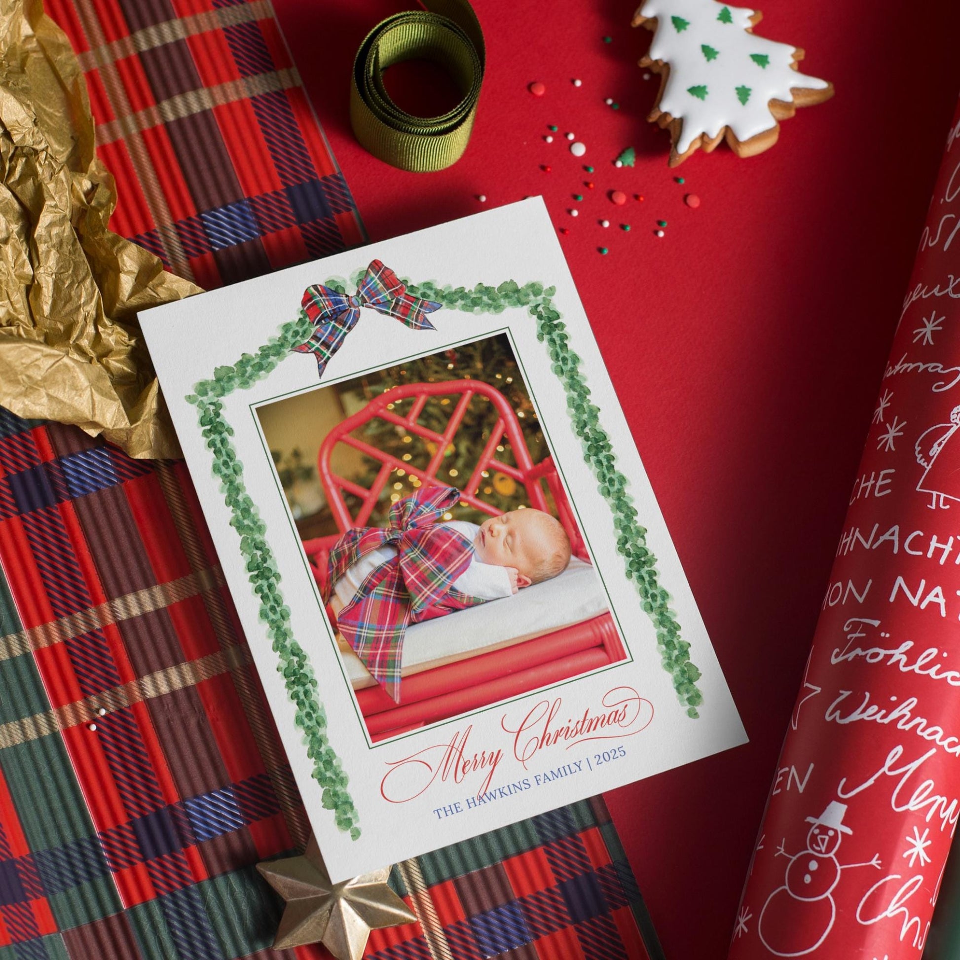 A Christmas card with a photo of a baby is placed on a red background, surrounded by festive decorations such as a ribbon, a cookie, and a snowman.