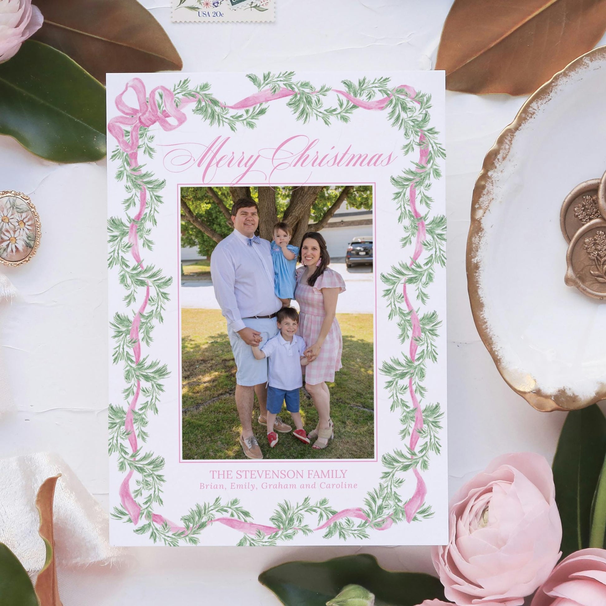 A family portrait of a man, woman, and child is displayed on a white card with a pink and green floral border. The card is surrounded by various decorative elements, including leaves, flowers, and a gold-colored shell.