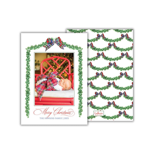 A Christmas card featuring a baby swaddled in a plaid blanket, with a red chair and a Christmas tree in the background. The card has a festive design with green and red bows, and the text &quot;Merry Christmas&quot; and &quot;The Hawkins Family (2023)&quot; is displayed.