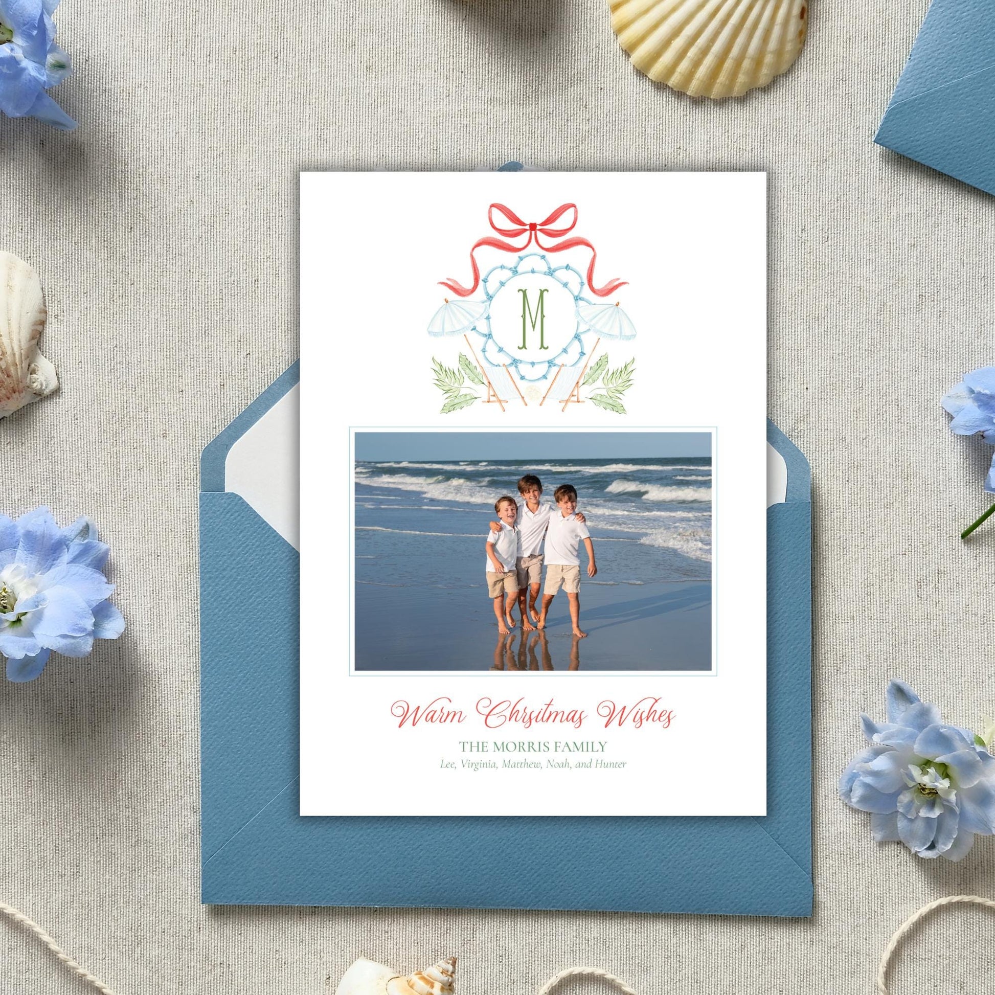 A holiday card featuring a photo of three children on a beach, with a ribbon and a letter M above the photo. The card is placed on a blue envelope, surrounded by seashells and blue flowers.
