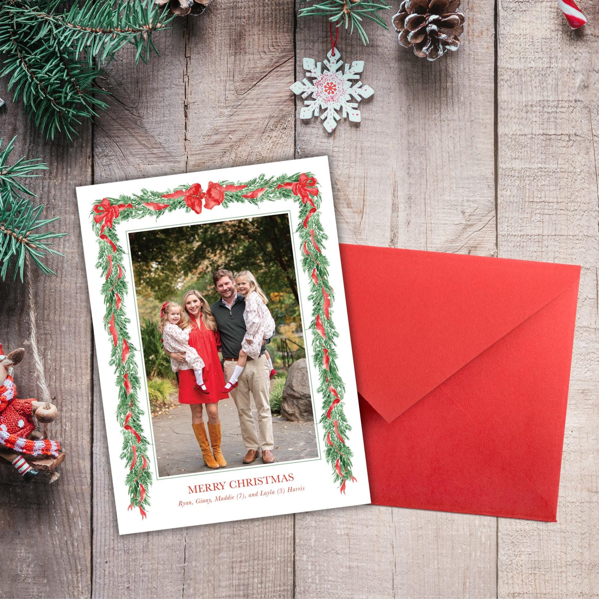 A Christmas card with a photo of a family is placed next to an envelope on a wooden surface, surrounded by Christmas decorations such as pine branches, pinecones, and a snowflake ornament.
