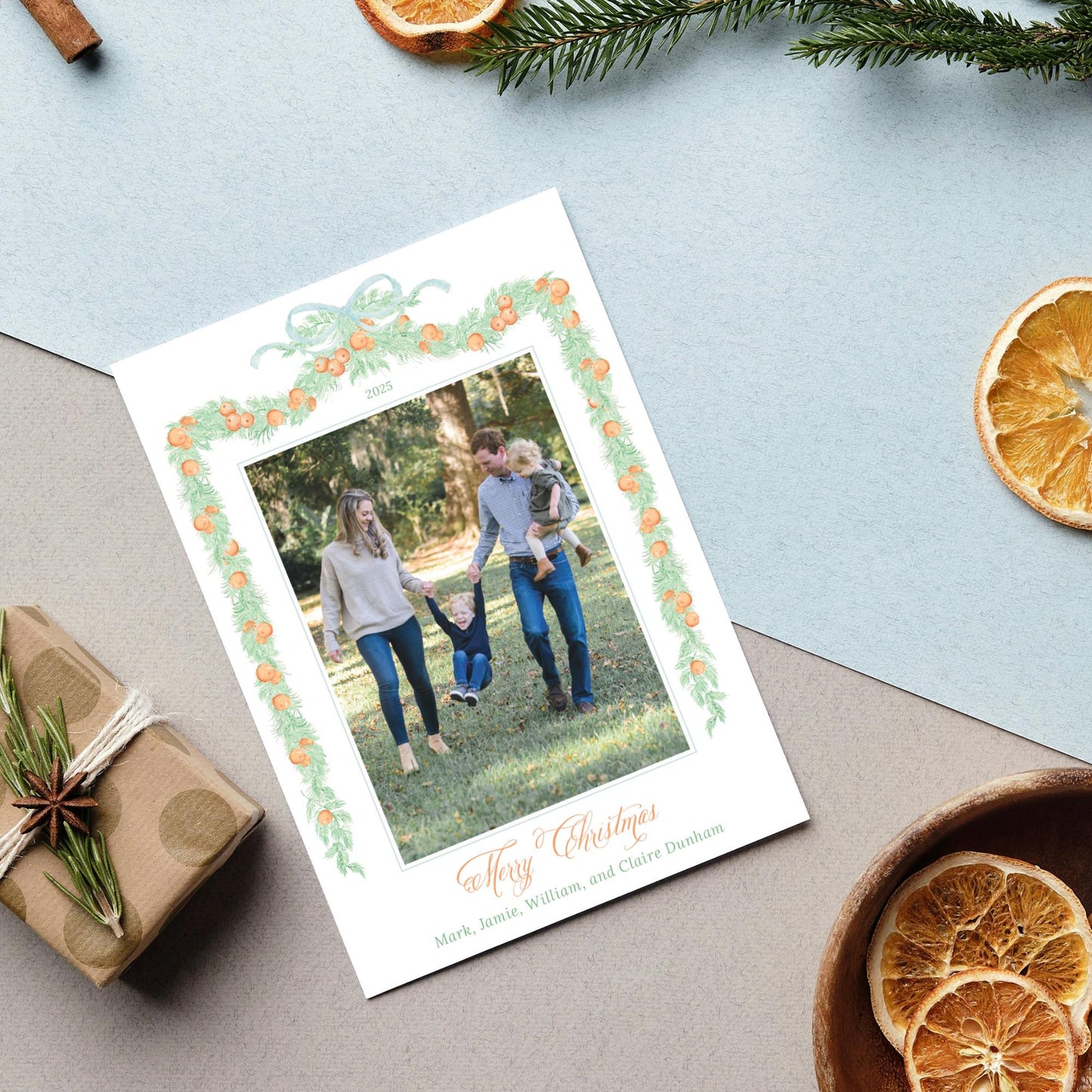 A family photo is displayed on a white card with a green border, surrounded by holiday decorations such as dried orange slices, pine branches, and a gift box.