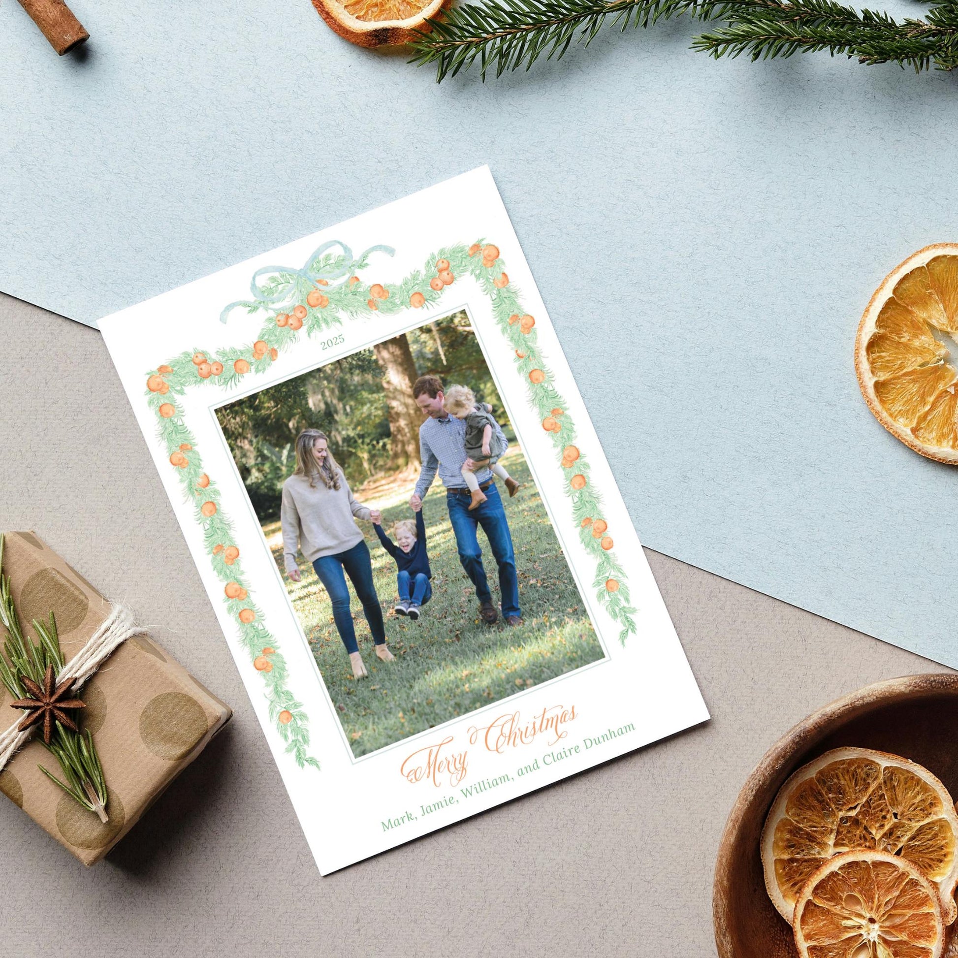 A family photo is displayed on a white card with a green border, surrounded by holiday decorations such as dried orange slices, pine branches, and a gift box.