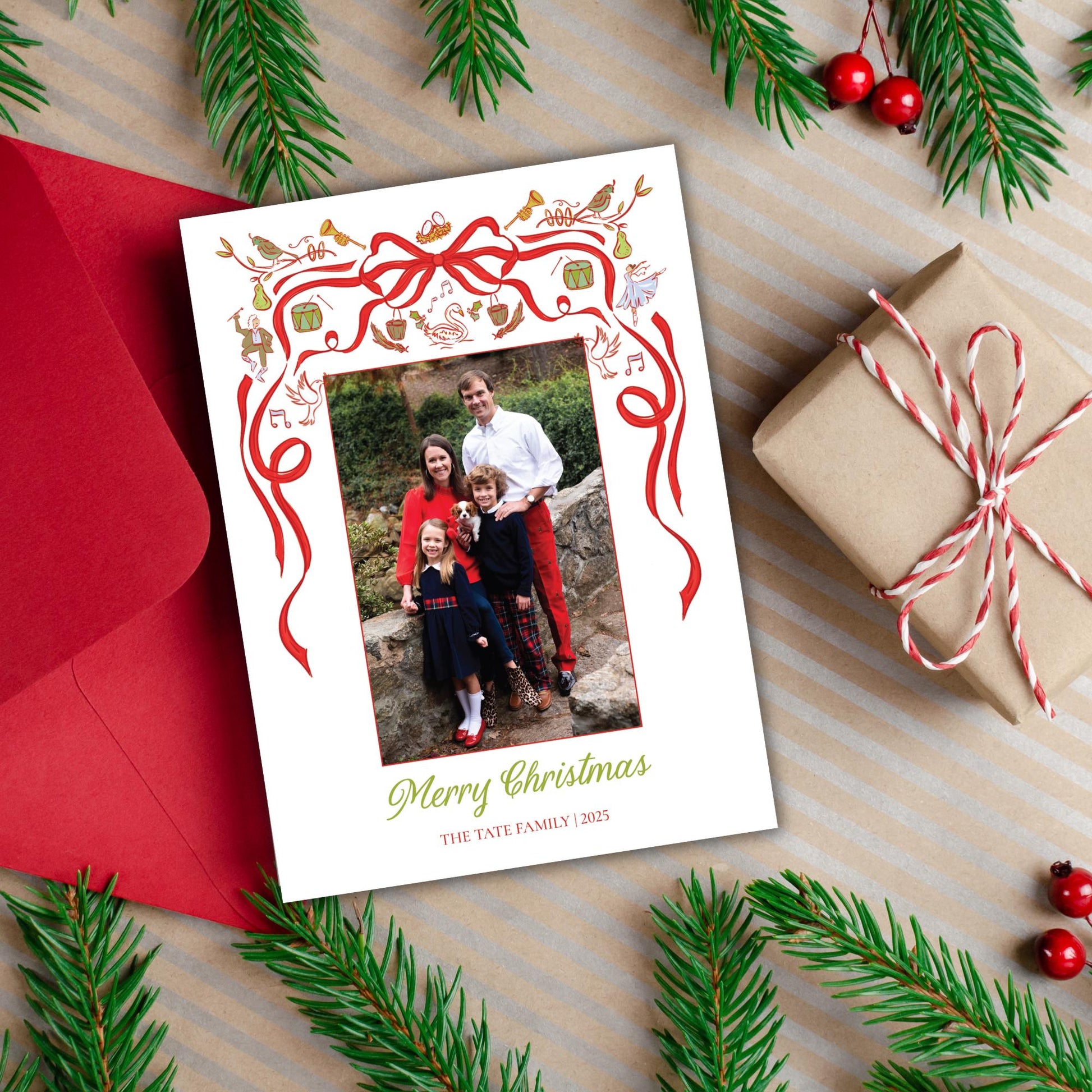 A Christmas card with a family photo is surrounded by holiday decorations, including pine branches, red and white ribbons, and a gift box.
