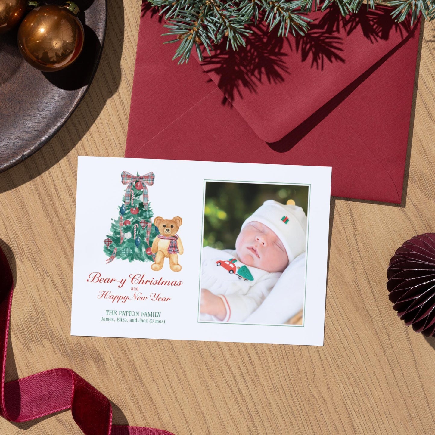 A white Christmas card with a photo of a baby and a teddy bear, along with a red envelope and Christmas decorations, including a tree and ornaments, on a wooden surface.
