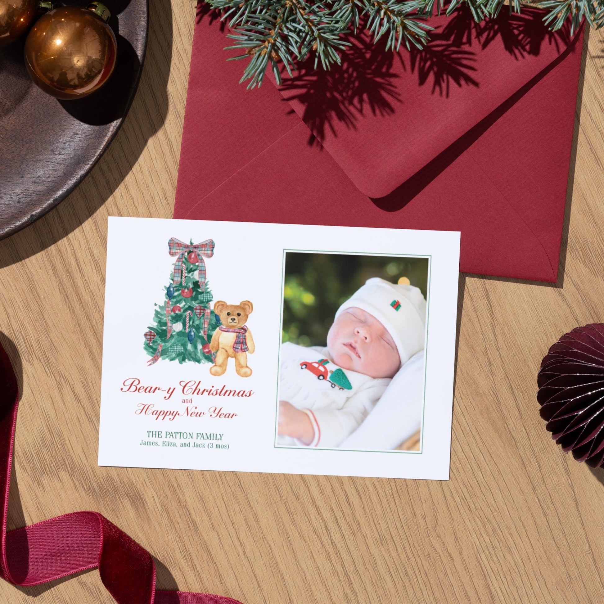 A white Christmas card with a photo of a baby and a teddy bear, along with a red envelope and Christmas decorations, including a tree and ornaments, on a wooden surface.