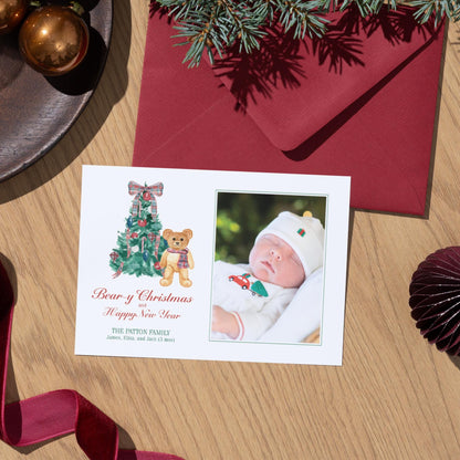 A white Christmas card with a photo of a baby and a teddy bear, along with a red envelope and Christmas decorations, including a tree and ornaments, on a wooden surface.