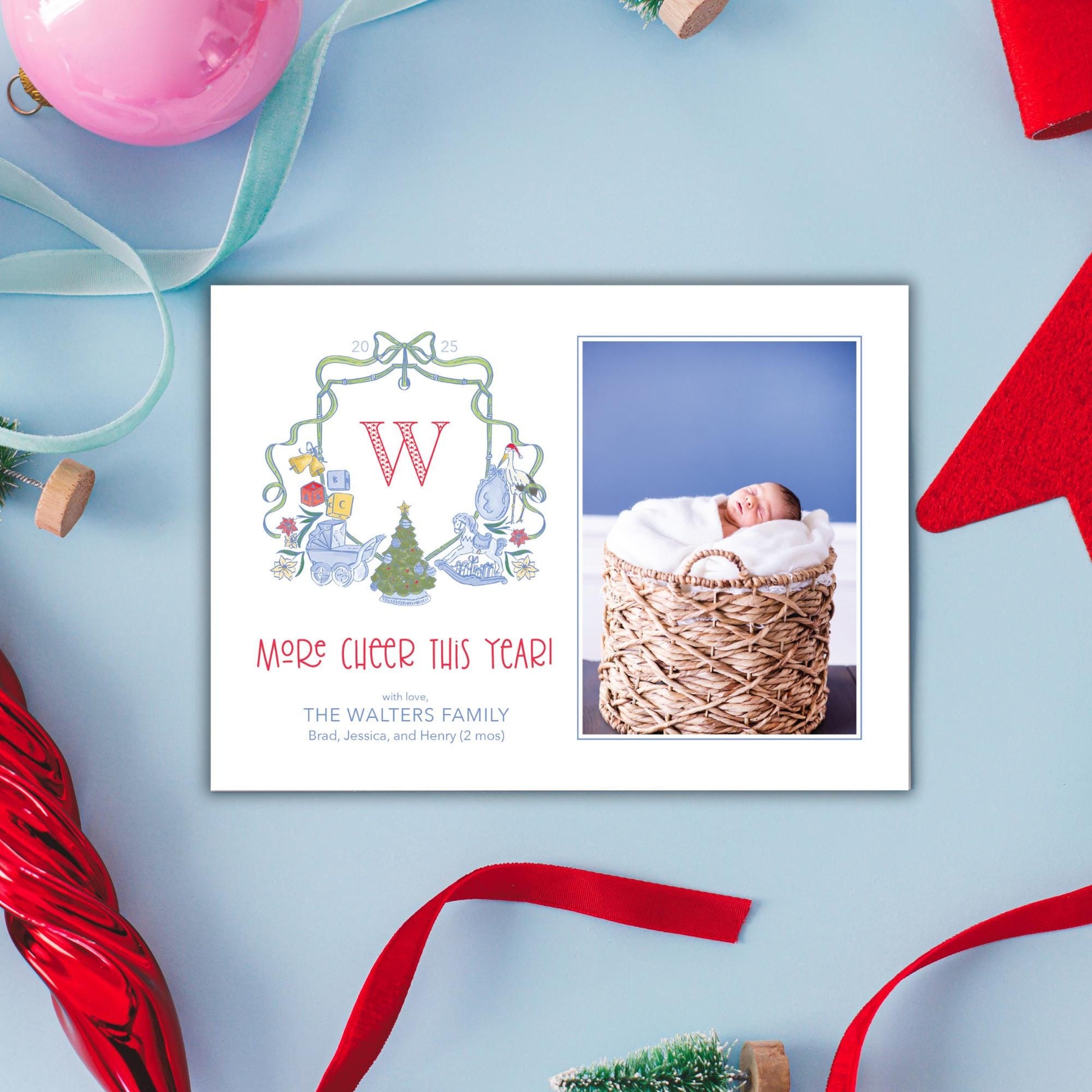 A white card with a photo of a baby in a basket, surrounded by Christmas decorations such as a red ribbon, a pink ornament, and a small Christmas tree.