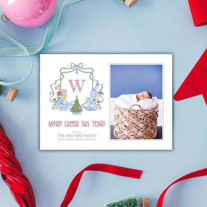 A white card with a photo of a baby in a basket, surrounded by Christmas decorations such as a red ribbon, a pink ornament, and a small Christmas tree.