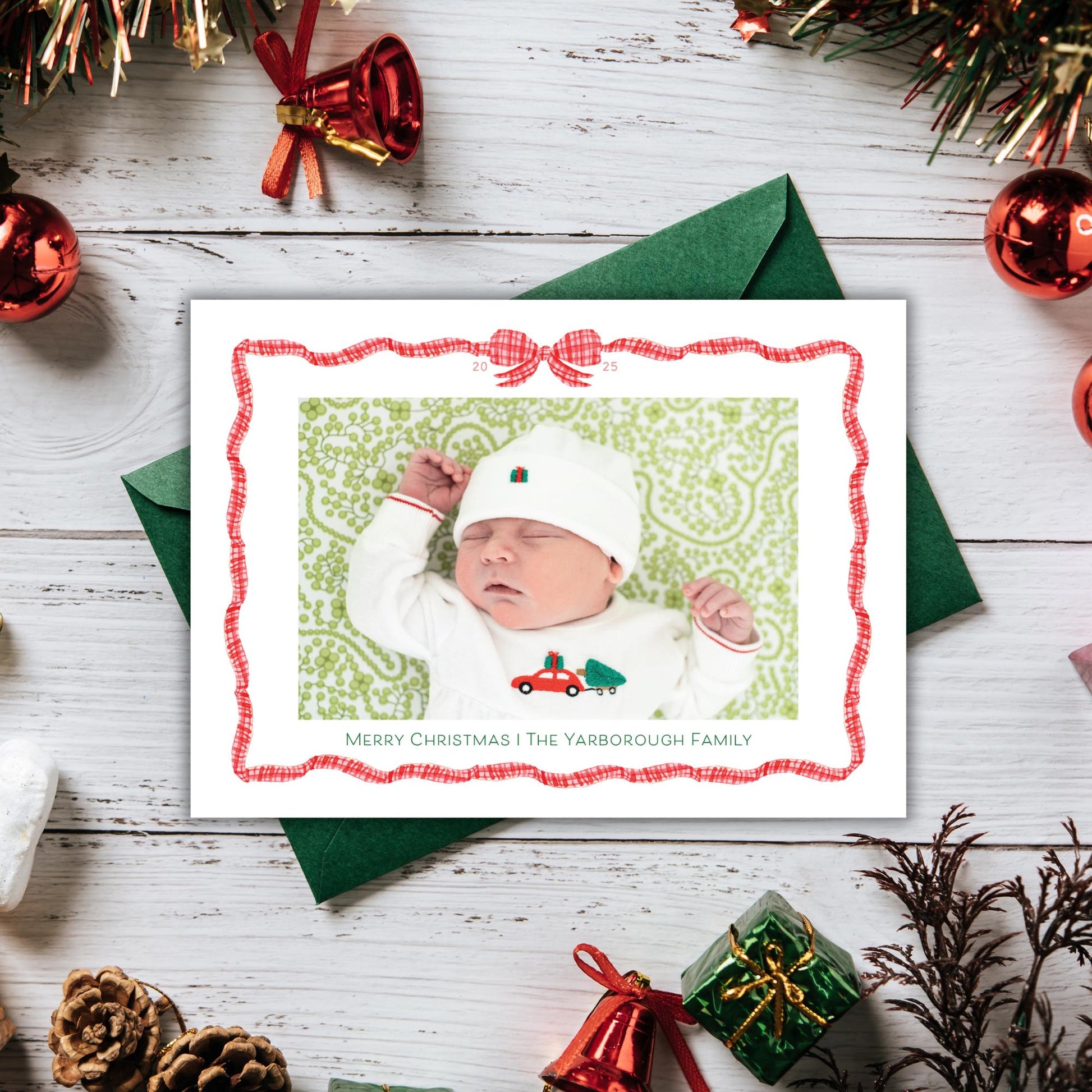 A Christmas card featuring a sleeping baby in a white onesie, surrounded by holiday decorations such as ornaments, tinsel, and a green envelope.