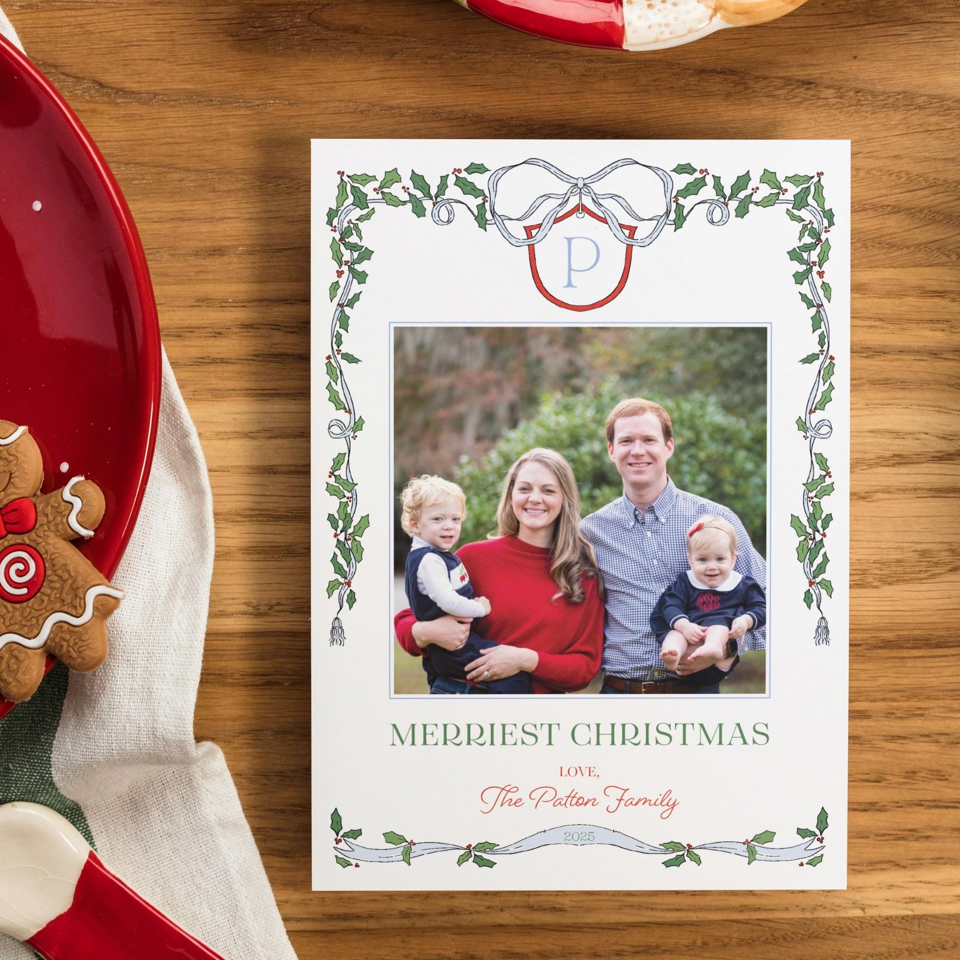A white Christmas card with a family photo of a man, woman, and two children, surrounded by a decorative border with holly leaves and a ribbon. The card is placed on a wooden surface, with a red plate and a gingerbread man nearby.