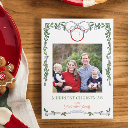 A white Christmas card with a family photo of a man, woman, and two children, surrounded by a decorative border with holly leaves and a ribbon. The card is placed on a wooden surface, with a red plate and a gingerbread man nearby.