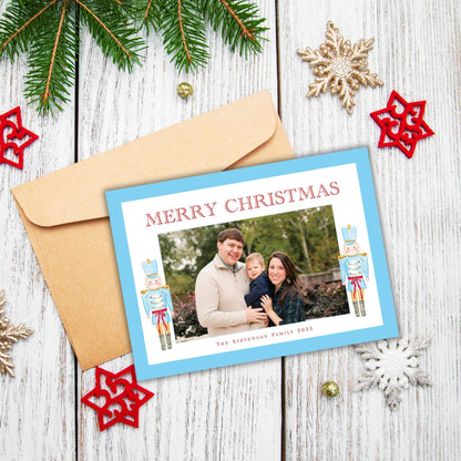 A Christmas card with a family photo and the text "Merry Christmas" is placed on a wooden surface, surrounded by Christmas decorations such as pine branches, star ornaments, and snowflakes.