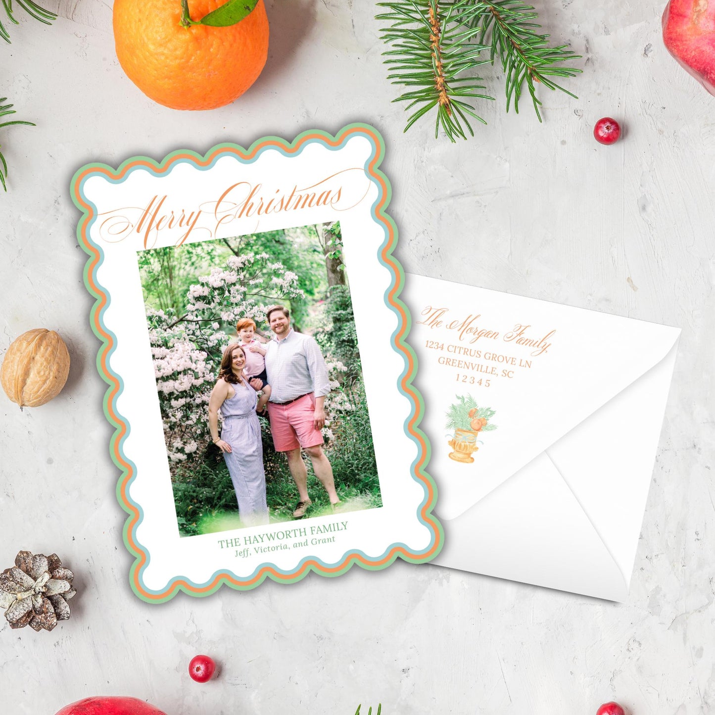 A Christmas card featuring a family photo of a man, woman, and child, along with the text &quot;Merry Christmas&quot; and &quot;The Hayworth Family Jeff, Victoria and Grant&quot;. The card is placed next to an envelope, and there are various holiday decorations such as an orange, pine branches, and berries on a white surface.