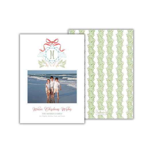 A holiday card featuring a family photo of three children on a beach, with a ribbon and a monogrammed letter &quot;M&quot; above them. The card has a green and white striped background and a message that reads &quot;Warm Christmas Wishes&quot; and &quot;The Morris family&quot;.