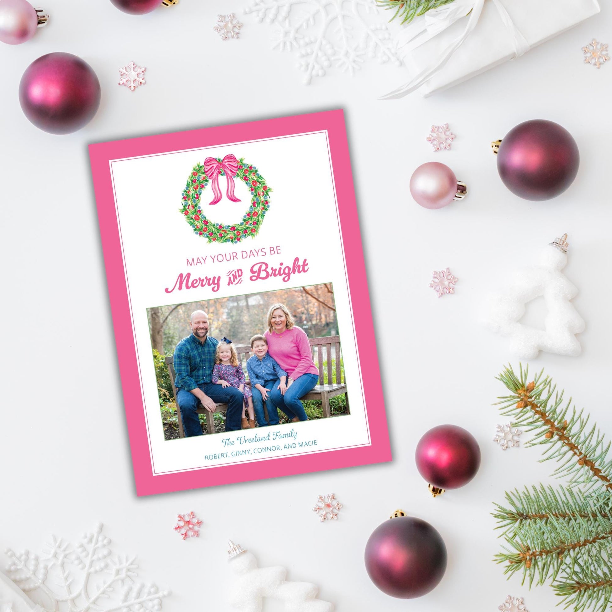 A pink greeting card with a family photo and the text &quot;May your days be Merry and Bright&quot; is surrounded by Christmas ornaments and pine branches on a white background.