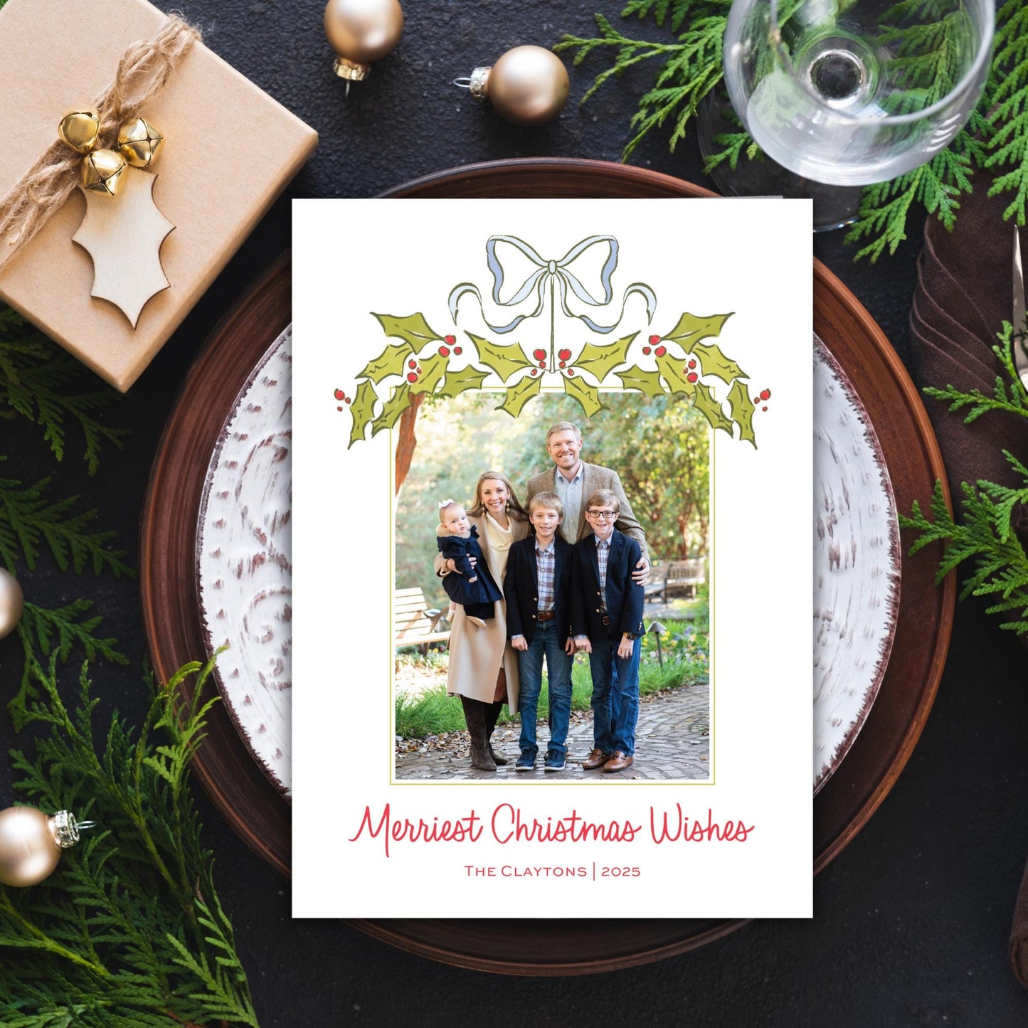 A Christmas-themed card with a family photo is displayed on a plate, surrounded by holiday decorations such as ornaments, pine branches, and a wine glass.