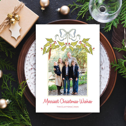 A Christmas-themed card with a family photo is displayed on a plate, surrounded by holiday decorations such as ornaments, pine branches, and a wine glass.