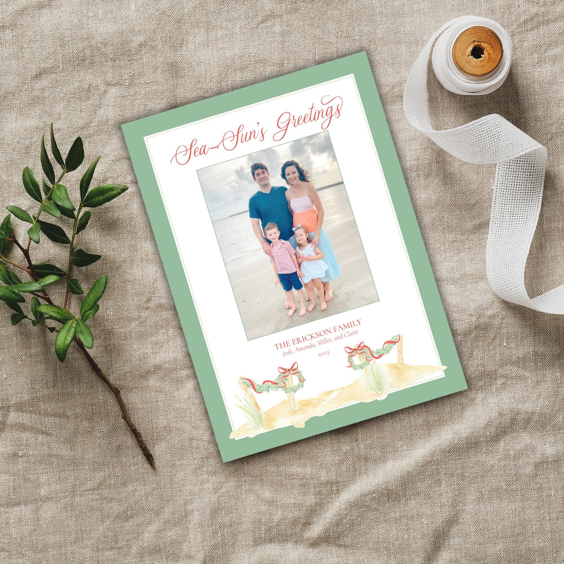 a green and white greeting card with a family photo on it, placed on a beige cloth. There is also a spool of ribbon and a small plant nearby.
