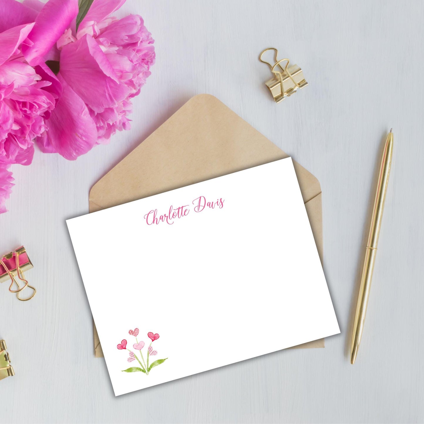 a blank personalized note card with a pink flower design, placed on a white surface alongside a gold pen, a gold binder clip, and a pink flower.